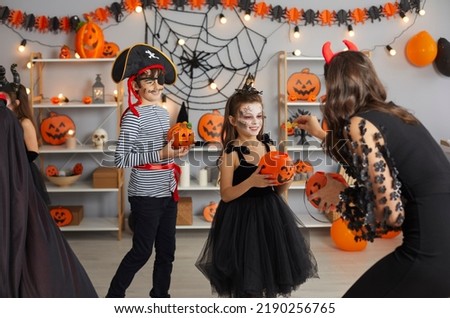 Young woman at Halloween party hands out candy to children who are dressed in spooky masquerade costumes. Woman throws sweets in pumpkin bucket to little girl in room with Halloween decor.