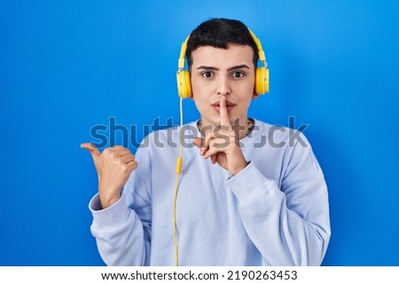 Non binary person listening to music using headphones asking to be quiet with finger on lips pointing with hand to the side. silence and secret concept. 