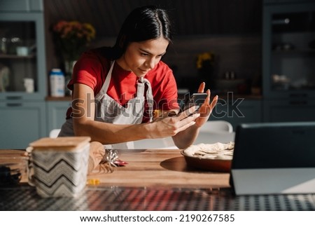 Young hispanic brunette woman wearing apron taking photo of meal while cooking in kitchen at home