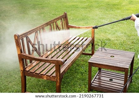 Close up view of person working cleaning pressure washing wooden garden furniture bench outdoors in summer. 