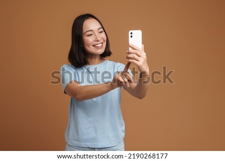 Young asian woman smiling while taking selfie on cellphone isolated over beige background