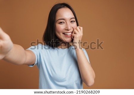 Young asian woman laughing while taking selfie photo isolated over beige background
