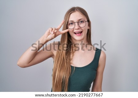 Young caucasian woman standing over white background doing peace symbol with fingers over face, smiling cheerful showing victory 
