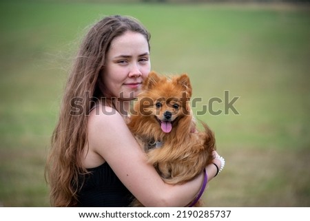 Portrait of a pretty young teenage girl  with long brown hair holding an orange fur fox nosed Pomeranian dog with grass in the background