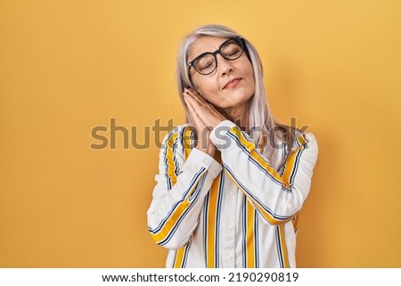 Middle age woman with grey hair standing over yellow background wearing glasses sleeping tired dreaming and posing with hands together while smiling with closed eyes. 