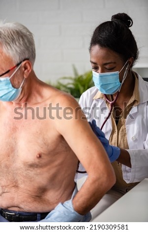 Female doctor is examining male elderly patient at clinic. Doctor and patient wearing a face mask.