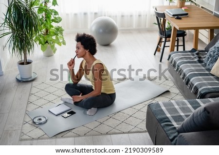 Young African-American woman writing in her notebook. Sitting at home by the window on a floor after exercise. Personal growth resolutions. Stay fit, loose weight. Royalty-Free Stock Photo #2190309589