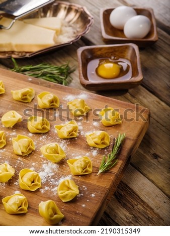 On a wooden cutting board are small homemade ravioli stuffed with minced meat, fragrant rosemary sprigs, cooking ingredients. Home recipes, family traditions.