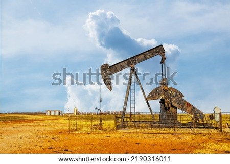 Pump jack oil gas well on red soil with storage tanks on horizon under dramatic sky