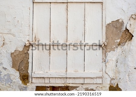White cracked wall with a peeling paint with a window closed with a painted white wooden shutters and locked with two locks