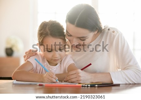 Loving happy young Caucasian mother and small daughter sit at table at home draw in album together. Smiling caring mom or nanny have fun play paint with little girl child. Hobby concept.