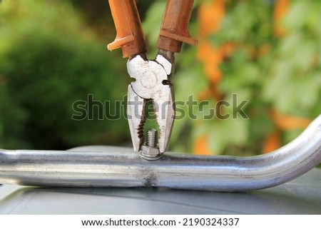 Metal pliers with brown plastic handles hold the nut vertically, tightening it. Aluminum pipe is screwed to the iron base with a bolt, nut and washer. Close-up view. Image with selective focus. Royalty-Free Stock Photo #2190324337
