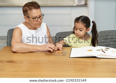 Child and grandmother making herbarium on dining wood table in home. Senior woman and little girl putting pressed flowers on the glass picture frame.