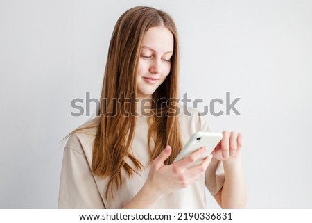 Lovely happy young woman holds modern cell phone, glad to get message from boyfriend, types feedback, checks balance online, wears t shirt stands over gray studio wall