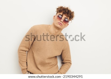 a handsome man in a beige sweatshirt stands on a light background wearing sunglasses
