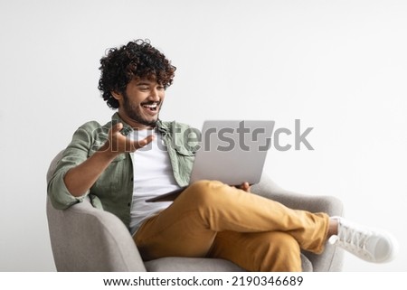 Relaxed handsome millennial eastern guy having video call with friend, sitting in armchair over white background, using computer, looking at laptop screen, smiling and gesturing, copy space