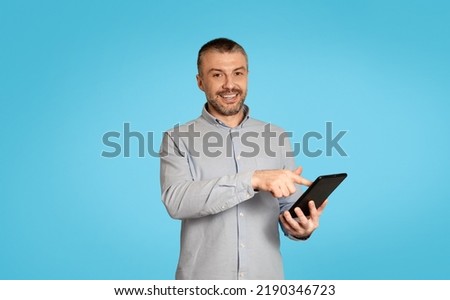 Happy Mature Male Using Digital Tablet Browsing Internet And Working Remotely Smiling To Camera Over Blue Studio Background. Freelancer Man Posing With Computer. Technology Concept