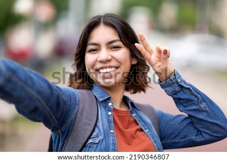 Cheerful Young Arab Woman Taking Selfie Outdoors, Joyful Beautiful Middle Eastern Female Student Showing Peace Gesture At Camera While Capturing Self-Portrait, Having Fun During Walk In City Royalty-Free Stock Photo #2190346807