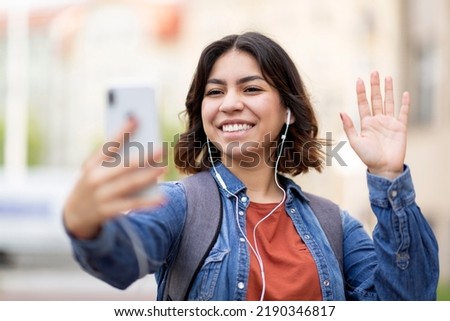 Happy young arab woman making video call via cellphone while standing outdoors, cheerful middle eastern female student wearing earphones waving hand at camera, gesturing hello, communicating online