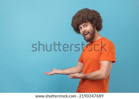 Please, take for free. Portrait of man with Afro hairstyle wearing orange T-shirt welcoming with wide open arms and smiling kindly, happy to embrace you. Indoor studio shot isolated on blue background