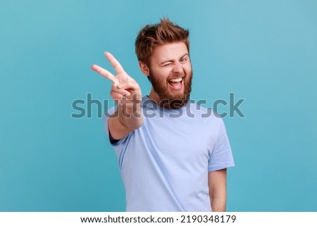 Portrait of happy optimistic bearded man doing victory gesture and winking playfully to camera, showing peace, v sign with double fingers. Indoor studio shot isolated on blue background. Royalty-Free Stock Photo #2190348179