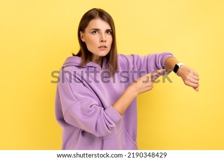 Portrait of bossy woman with dark hair pointing wrist watch and looking annoyed displeased, showing clock to hurry up, wearing purple hoodie. Indoor studio shot isolated on yellow background. Royalty-Free Stock Photo #2190348429