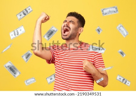 Money rain, winner and rich. Ecstatic happy celebrating motivated man standing with raised fists and shouting for joy, winner excited for success. dollars falling. indoor isolated on yellow background