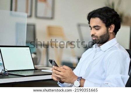 Indian young business man company worker, professional employee, businessman executive holding smartphone using cellphone mobile corporate apps texting, checking financial data at work in office.