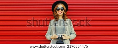 Portrait of young woman photographer with film camera wearing knitted sweater cardigan, black round hat on red background