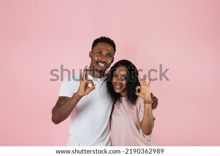 Photo below belt of cheerful people of exotic appearance on isolated nacre pink wall. African female with her arms on waist dressed in pink T-shirt and jeans while guy in white t-shirt showing sign