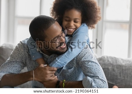 Close up smiling African American father piggy backing adorable daughter, sitting on cozy couch at home, overjoyed dad wearing glasses and little girl hugging, enjoying leisure time together Royalty-Free Stock Photo #2190364151
