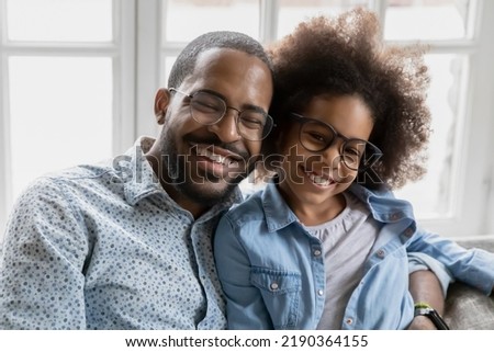 Head shot portrait happy African American father and adorable daughter wearing glasses hugging, looking at camera, loving dad and little girl child bonding, embracing, posing for photo together