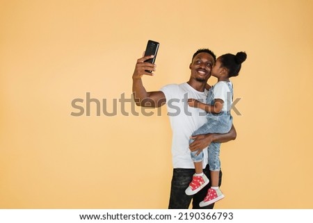 Cheerful african cute little girl kissing her father in casual clothes isolated on yellow background studio portrait. People lifestyle concept. Mock up copy space. Doing selfie shot on mobile phone.