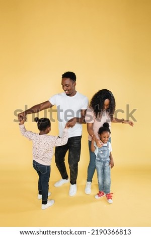 Portrait of happy family in colorful stylish outfits, young African woman mom, her husband and two cute little kids girls, having fun, playing and dancing together, on isolated yellow background.