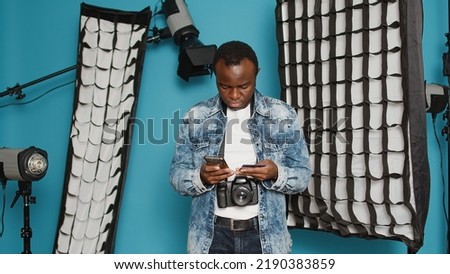 Photographer with camera equipment in studio doing online shopping, using smartphone and credit card to make purchase from retail store website. Having professional photography tools.