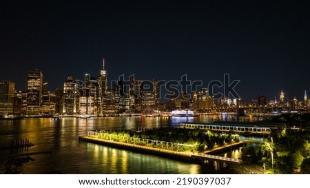 An aerial view of Brooklyn Bridge Park at night with lower Manhattan in the background, reflecting lights onto the calm and reflective East River.