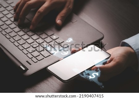 Hand holding smartphone and using computer laptop with virtual global and currency sign for currency exchange and global trade forex concept. 