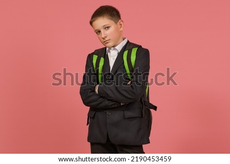 a serious schoolboy in a black suit with a briefcase crossed his arms on his chest on a colored background
