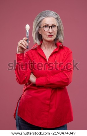 Woman in eyeglasses woth a face brush in hnd looking thoughtful