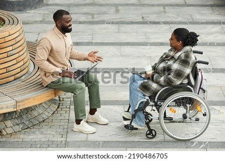 Full length shot of adult black couple with woman in wheelchair chatting outdoors in city setting and enjoying coffee
