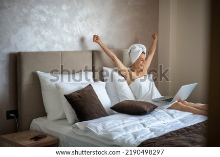 Young cheerful happy woman sitting on bed wrapped in bath towels using laptop with arms outstretched