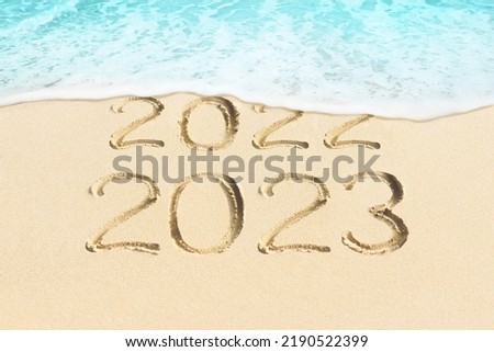 New Year concept photo. Text 2023 and 2022 handwritten in sand surface. Blue ocean wave washing away numbers on beach.  Royalty-Free Stock Photo #2190522399