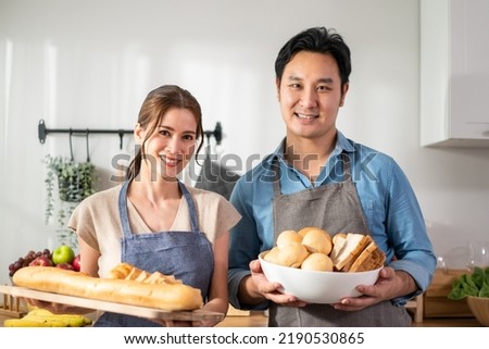 Portrait of Asian young couple hold a bowl of bread and look at camera. Attractive man and woman wear apron, spend time cooking foods breakfast for health care in house. Diet and Healthy food concept.