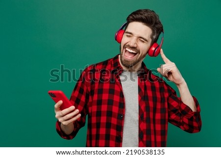 Young happy smiling cool man he 20s in red shirt grey t-shirt headphones listen to music dance hold mobile cell phone isolated on plain dark green background studio portrait. People lifestyle concept