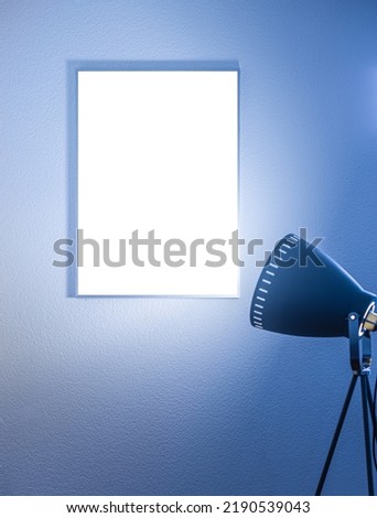 Blank vertical poster in a frame on the wall illuminated by a tripod floor lamp