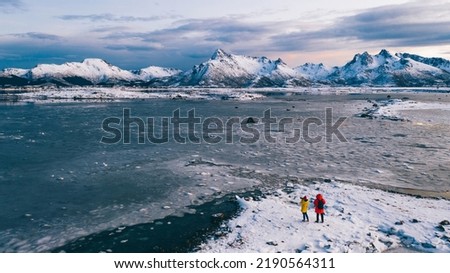 Aerial view from drone of male and female photographers in expedition walking on frozen shore of lake in Scandinavia, wanderlust couple of bloggers with cameras taking photo of scenery winter nature