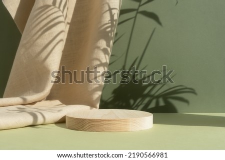 Wooden cylindrical podium, pedestal on a green background with beige curtain and a shadow of palm leaves. Scene for the presentation of a cosmetic product. Empty showcase