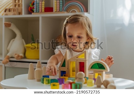 A little girl playing with wooden blocks on the table in playroom.  Educational game for baby and toddler in modern nursery. The kid builds a tower from wooden rainbow stacking blocks. Royalty-Free Stock Photo #2190572735