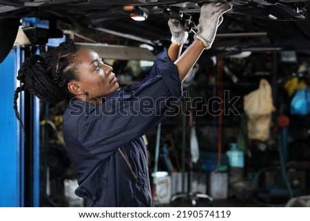 Woman technician car mechanic in uniform checking maintenance a lifted car service at repair garage station. Worker using flashlight and wrench fixing breakdown vehicle. Car repair service concept.