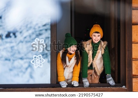 Two happy fun children are playing by the open window in winter time. Children are dressed in warm winter knitted clothes. Christmas decorations on the window.